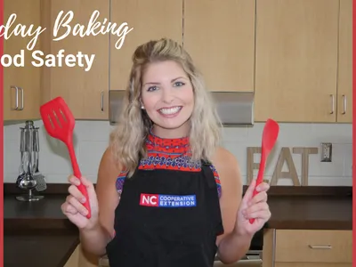 Person in apron holding red spatulas in a kitchen; text: "Holiday Baking", "Food Safety", "NC Cooperative Extension"