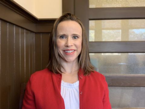 Woman facing camera, wearing red jacket, in front of wooden paneling and frosted glass