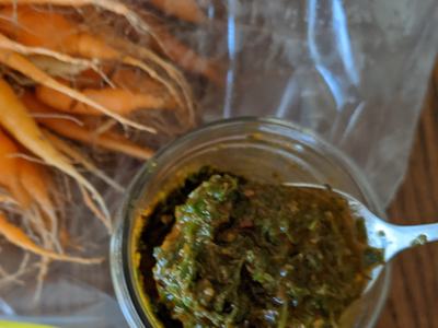 Jar of green herb pesto on a spoon, small carrots in a plastic bag beside it