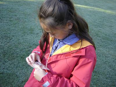 Child in red jacket inserting earphones into a portable music player while standing on grass
