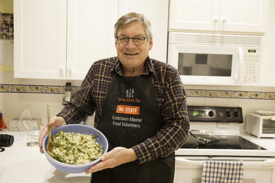Man holding bowl of rice and greens in kitchen; apron reads "NC STATE Extension Master Food Volunteers"
