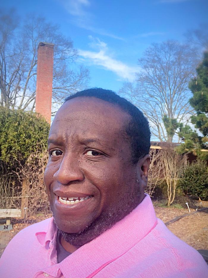 Person wearing a pink polo shirt outdoors, with trees and a brick chimney in the background