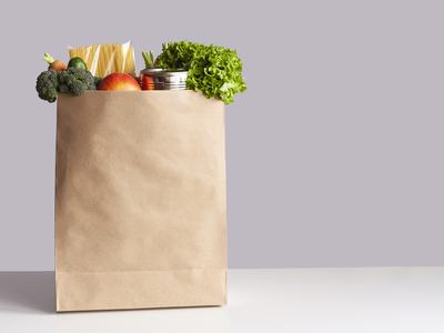 Various grocery items in paper bag on white table opposite gray wall. Bag of food with fresh vegetables, fruits, pasta and canned goods.