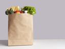 Various grocery items in paper bag on white table opposite gray wall. Bag of food with fresh vegetables, fruits, pasta and canned goods.