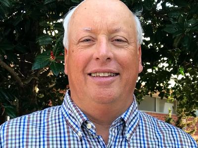 Bald man in checked button-down shirt standing before a leafy tree with red berries
