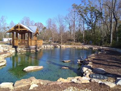 Wooden gazebo on a stone-lined clear pond with surrounding trees and a path