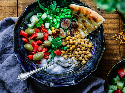 Bowl with yogurt topped with roasted chickpeas, olives, cucumber, peas, figs, pita wedge and spoon