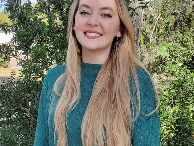 Young woman with long blonde hair in teal sweater standing outdoors by trees