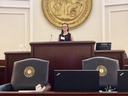 Person standing at courtroom lectern with large circular state seal on wall behind