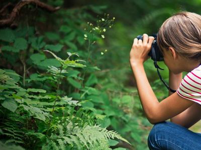 Young girl crouching with binoculars observing plants on a forest trail