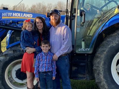 Family of four (parents and two children) standing beside blue New Holland tractor