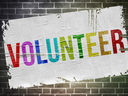 Poster on brick wall reading "VOLUNTEER" in rainbow-colored letters