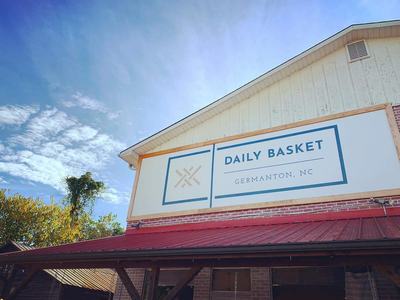 Storefront sign reading "DAILY BASKET" "GERMANTON, NC" mounted on a building under a blue sky
