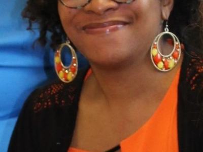 Person smiling, wearing glasses, large colorful hoop earrings and an orange top.