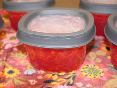 Small clear plastic container with gray lid filled with red jam on a floral cloth