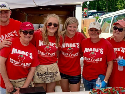 Six people in red "Mabe's Berry Farm" shirts selling boxes of strawberries at a stall; sign "STRAWBERRIES"