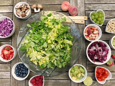 bowl of lettuce with surrounding bowls of fixings for salad like blueberries, strawberries, grapes, mushrooms, avocado, and tomatoes