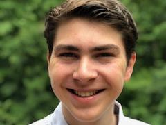 Young man smiling, wearing a white collared shirt, blurred green foliage background.