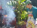 Young man in green cap and tie-dye apron standing beside a smoking charcoal grill