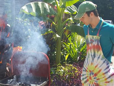 Young man in green cap and tie-dye apron standing beside a smoking charcoal grill