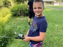 Young boy holding a fishing rod on a grassy bank, another person standing in background.