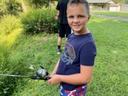 Young boy holding a fishing rod on a grassy bank, another person standing in background.