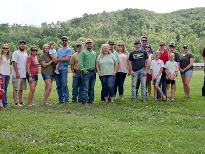 Large group of adults and children standing in a grassy field with wooded hills behind