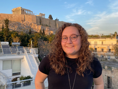 Person with curly hair and glasses standing on a rooftop with the Acropolis (Parthenon) in background