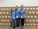Three women in blue shirts standing in front of stacked cardboard boxes