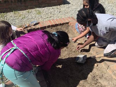 Four children kneeling over a brick garden bed digging and planting with trowels.