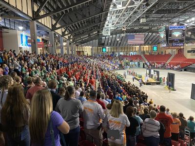 Crowd standing in arena facing a central stage on the floor beneath a hanging American flag