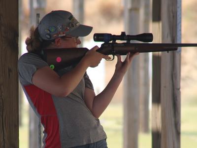 Shooter aiming a scoped rifle at an outdoor firing range, resting the stock against their shoulder