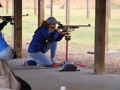 Shooter kneeling at an outdoor range aiming a rifle supported on a red shooting rest