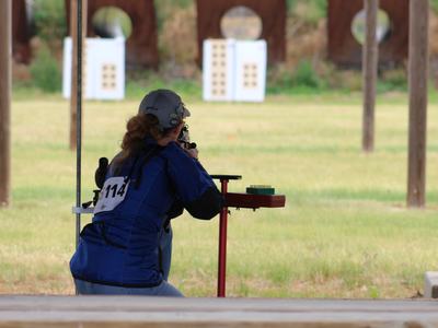 Shooter in blue jacket kneeling and aiming a rifle at targets on a shooting range, bib 114