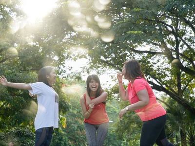 Three women standing and talking in a sunlit park surrounded by trees