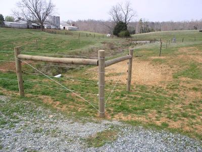 Wooden fence corner with barbed wire beside rural gravel driveway and pasture
