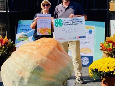Two people holding 2021 Nash County 4-H sign showing pumpkin weight 910.0 behind giant pumpkin