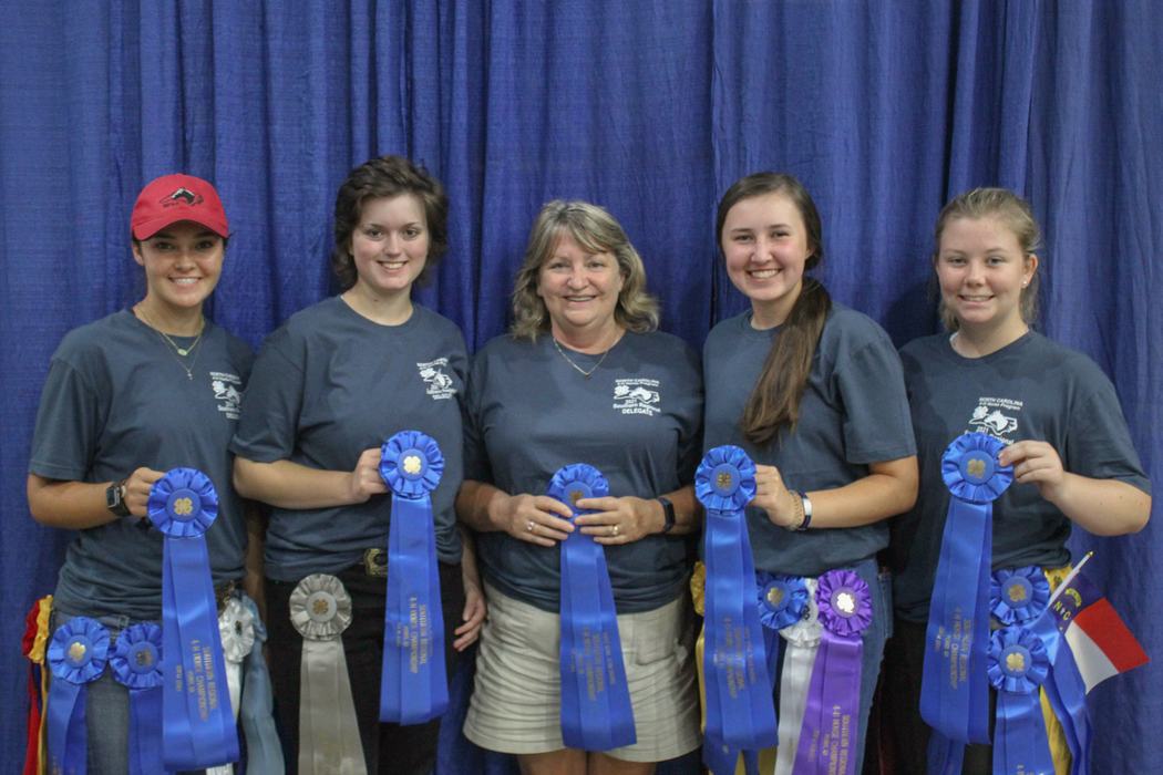 Five people standing side-by-side holding blue prize ribbons against a blue curtain