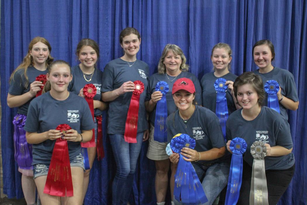 Nine people holding red, blue, purple, and gray award ribbons in front of a blue curtain