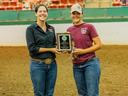 Two women standing in an indoor dirt arena holding an award plaque together