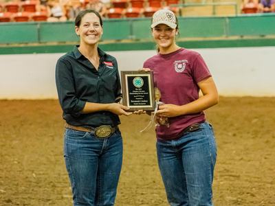 Two women standing in an indoor dirt arena holding an award plaque together