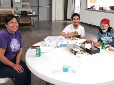 Three people at round table eating pizza, soda cans, pizza boxes; hoodie reads "VCEC"
