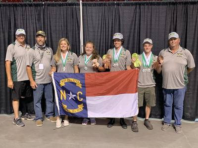 Eight-person team wearing matching shirts holding North Carolina flag reading "N C" and medals/ribbons