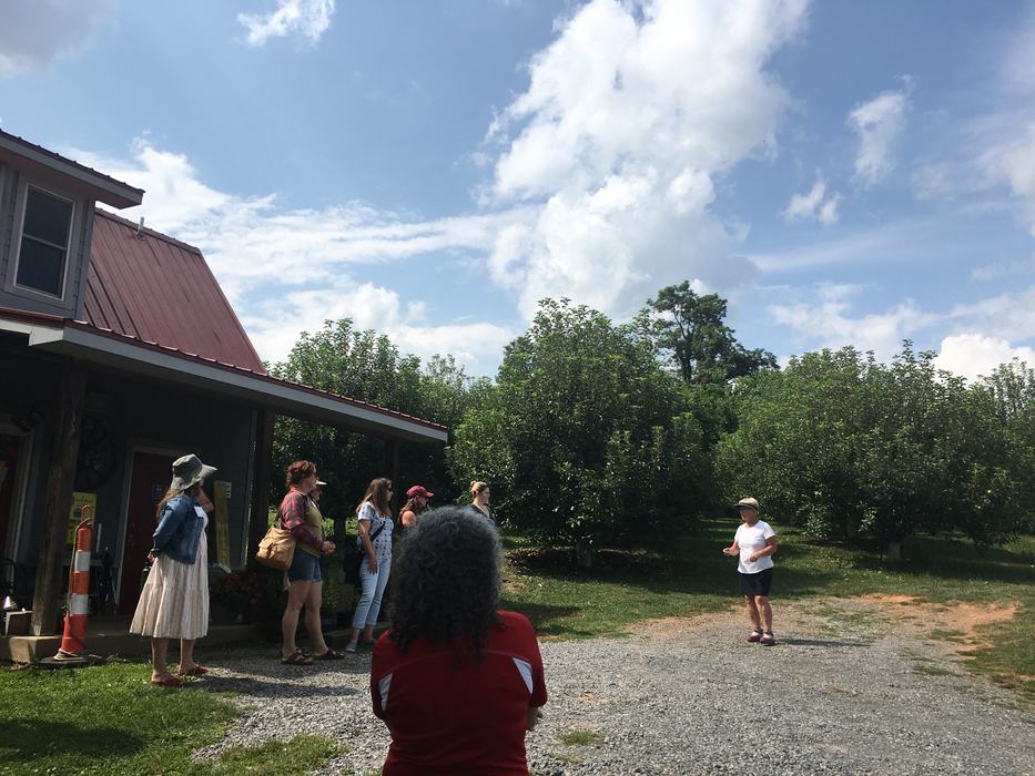 A farmer addresses a group of visitors with a backdrop of an apple orchard, blue skies and a small building. The farmer is discussing her farm's origin story and since development. 