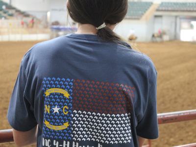 Person from behind wearing NC 4‑H Horse Program T-shirt in indoor horse arena