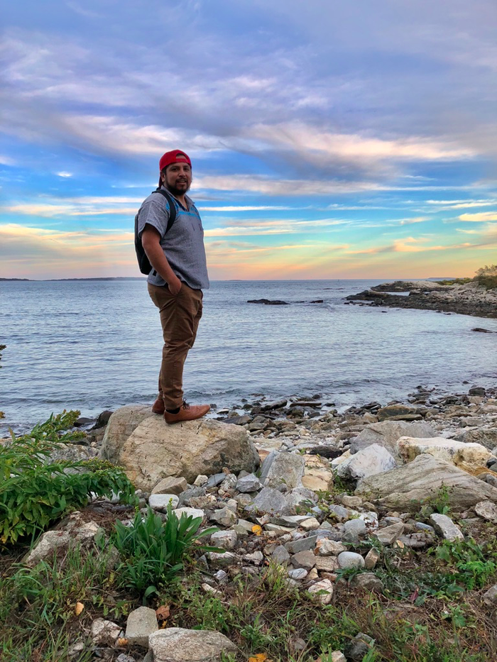 Man wearing red cap and backpack standing on a rock by a rocky shoreline at sunset