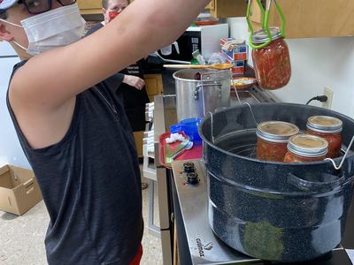 Masked person using tongs to lift a sealed jar from a water-bath canner with jars inside