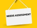 White hanging sign pinned with red pushpin reading "NEEDS ASSESSMENT" on yellow background