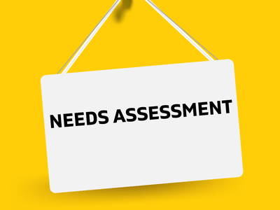 White hanging sign pinned with red pushpin reading "NEEDS ASSESSMENT" on yellow background