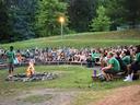About 100 youth sitting on bleachers around a campfire watching a man talk